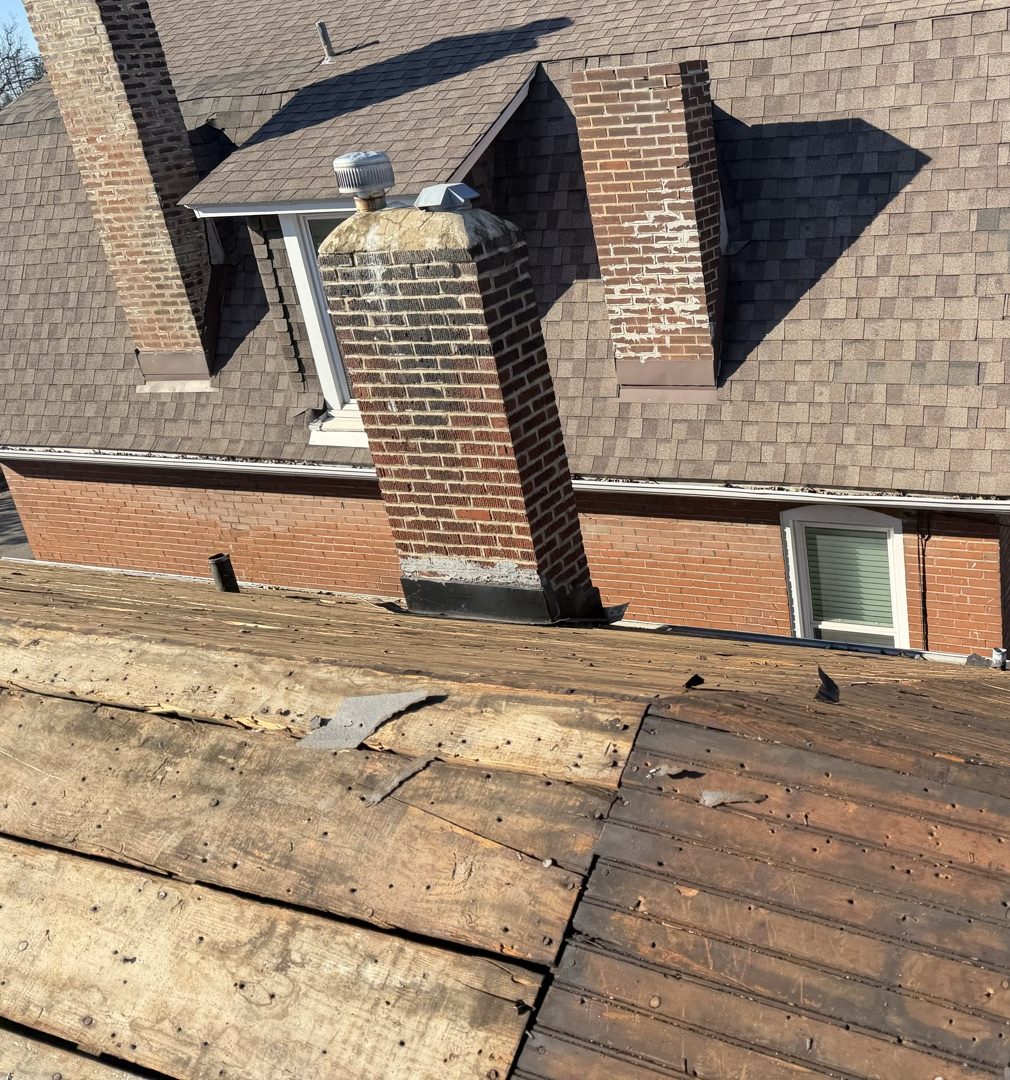An older two storey red brick house photographed from its stripped lower roof plane, the dark weathered plank decking running across the foreground with old nails still in the boards, while the main roof behind carries worn brown shingles, two brick chimneys with spalled faces and patched mortar, and a dormer with a metal turbine vent