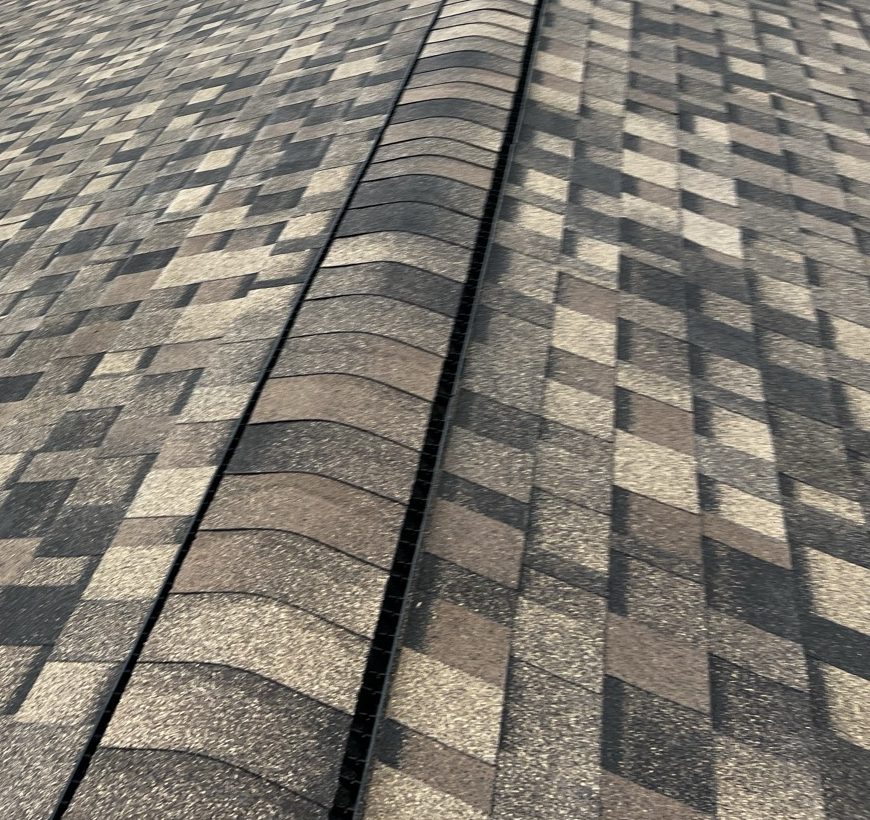 Close view along the ridge of a newly laid brown, tan and charcoal multi tone architectural shingle roof, with ridge cap shingles laid in an overlapping run over a dark ventilation strip, and the shingle courses of both planes running away below the cap