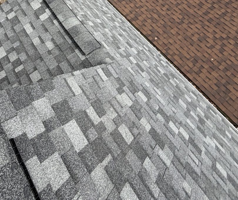 Looking down at the hip corner of a newly laid grey and charcoal architectural shingle roof, capped with ridge cap shingles over a dark vent strip that runs up the hip, with the weathered brown roof of the adjoining structure along the right edge