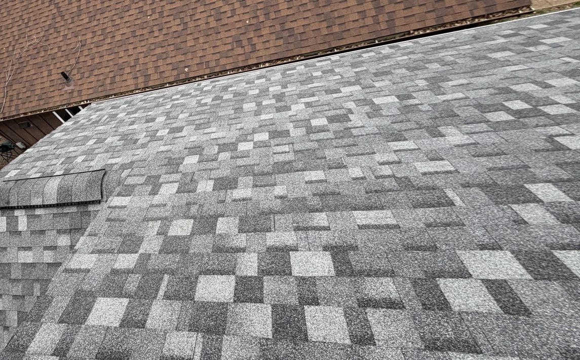 Seen from above, a newly laid grey and charcoal architectural shingle plane with a short capped ridge and a dark vent strip at the left, butting against the eave and gutter of the adjoining garage roof, which is still covered in old brown shingles