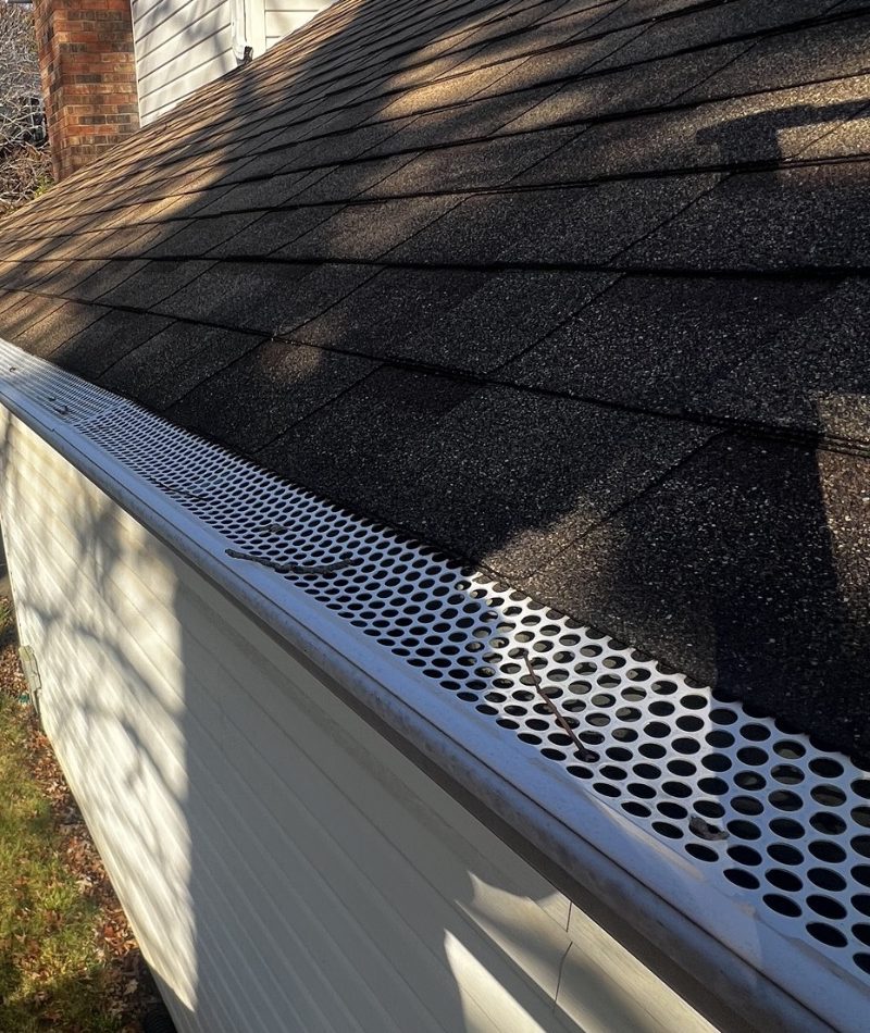 Looking along the eave of a dark charcoal shingle roof with a white perforated metal gutter guard, punched with round holes, fitted the length of the gutter. A bare twig and a dark scrap of debris rest on top of the perforations, white vinyl siding runs below the gutter, a red brick chimney stands at the upper left, and a strip of lawn with fallen autumn leaves shows at the lower left