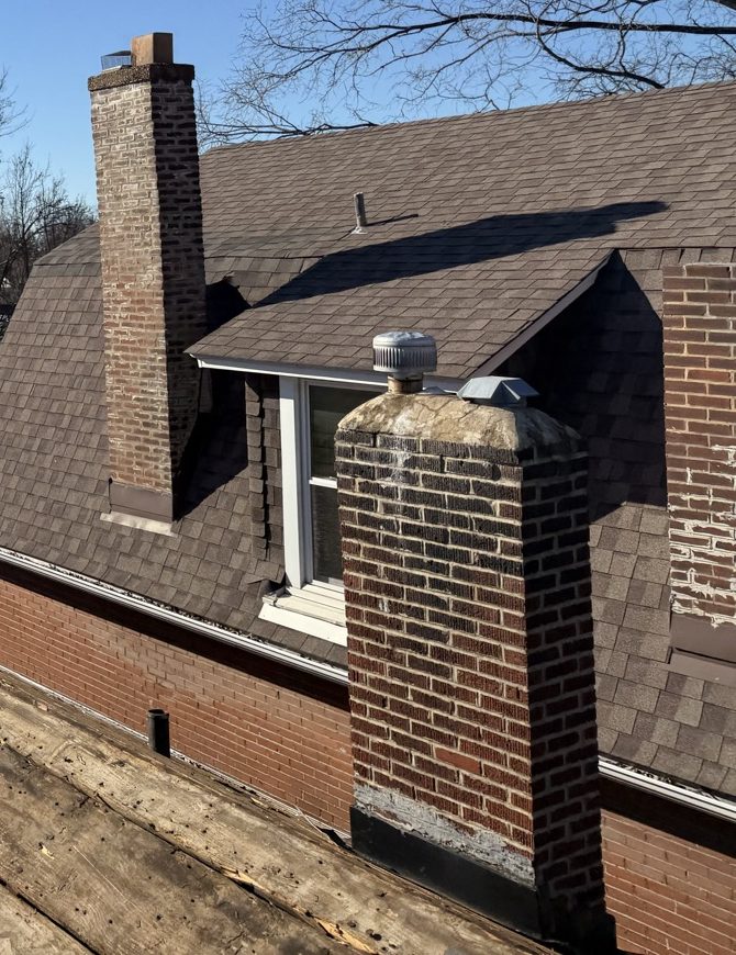 Bare plank decking running across the foreground of a stripped roof plane, and behind it the same house's brown shingled main roof with a tall brick chimney, a squat second chimney carrying a metal cap and a turbine vent, a dormer window and a red brick wall, under bare winter branches and a clear blue sky