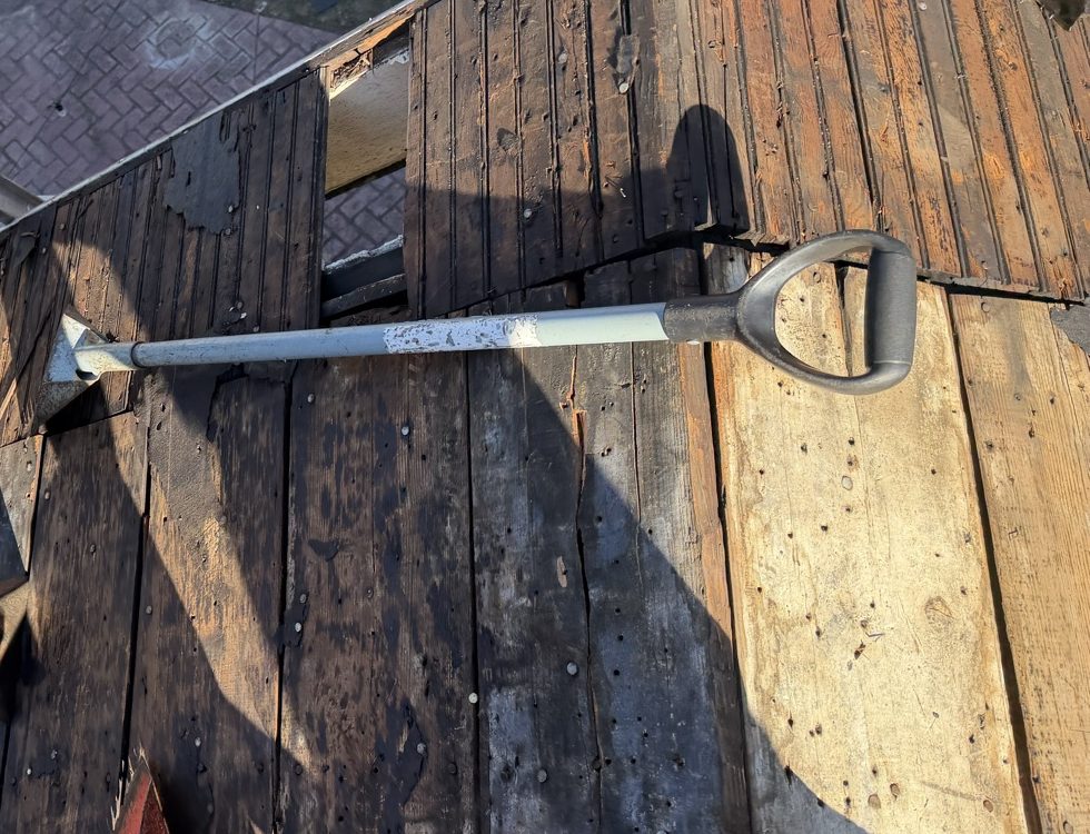Overhead view of stripped plank decking with torn felt still clinging to the boards, an aluminium roofing shovel with a D shaped handle laid across the run, a red spade blade at the corner of the frame, and the hip of the decking falling away toward a brick paver patio below