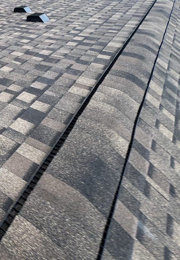 A newly laid brown, tan and charcoal multi tone architectural shingle roof with two low black box vents set near the top of the plane and the capped ridge running down the right of the frame over a dark ventilation strip